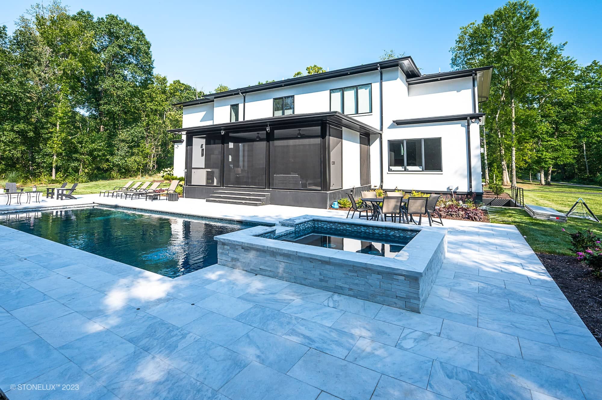 Afyon Elegance: Modern white two-story marvel with black accents features large windows, a spacious patio, an in-ground pool, and a square hot tub surrounded by trees and lounge chairs under a clear blue sky.