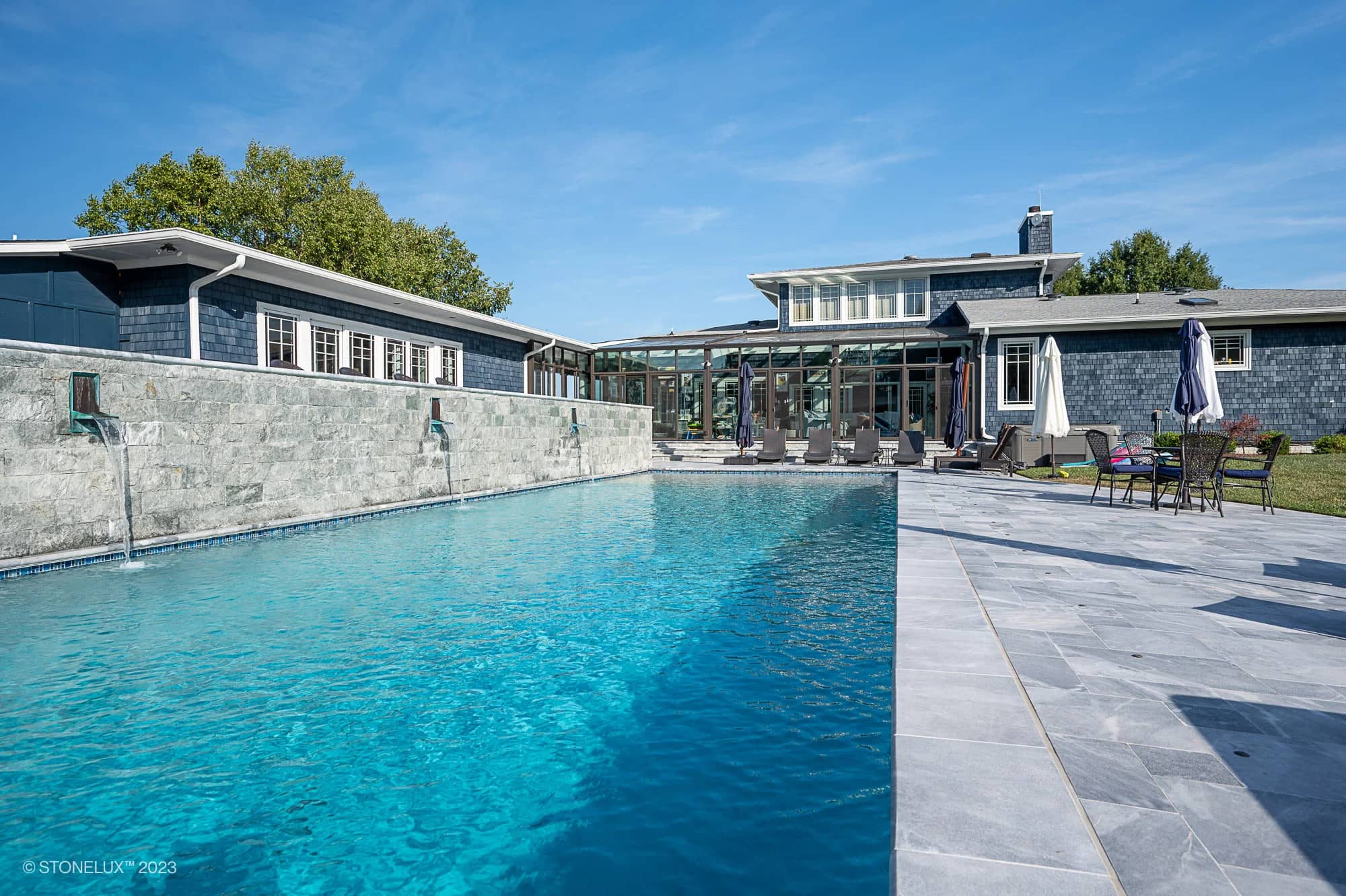 Modern outdoor swimming pool, surrounded by grey stone tiles. Blue Haven: The Grey and Blue Sandblasted Marble Pool Project features a stunning stone wall with water features, lounge chairs, patio furniture, and a large blue-sided house.