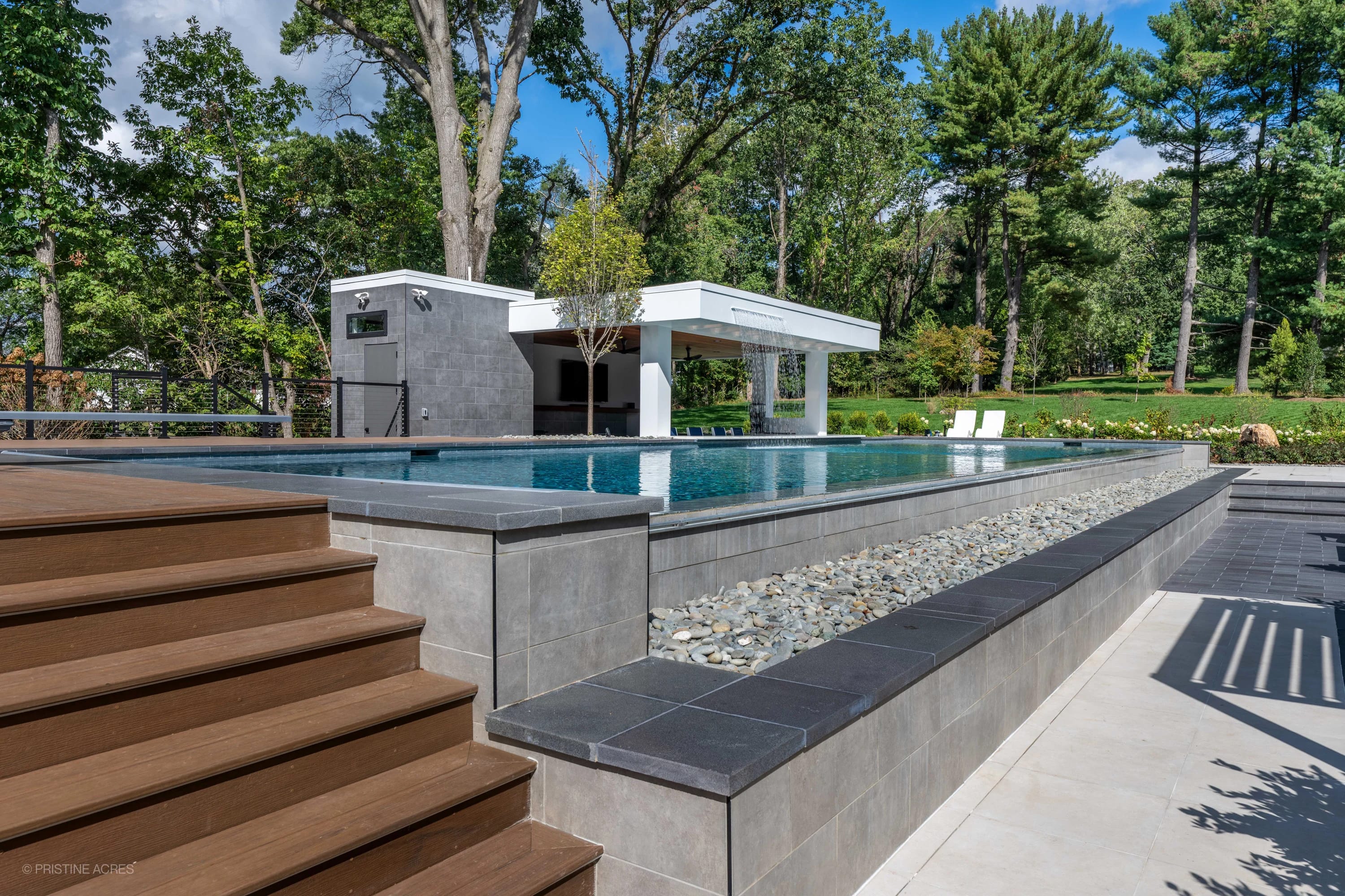 Modern McLean Pool Project in McLean VA showcases a sleek design using Black Basalt and Nexus White Porcelain Pavers. The luxurious pool features a waterfall, surrounded by lush greenery and white lounge chairs, creating a serene backyard oasis.