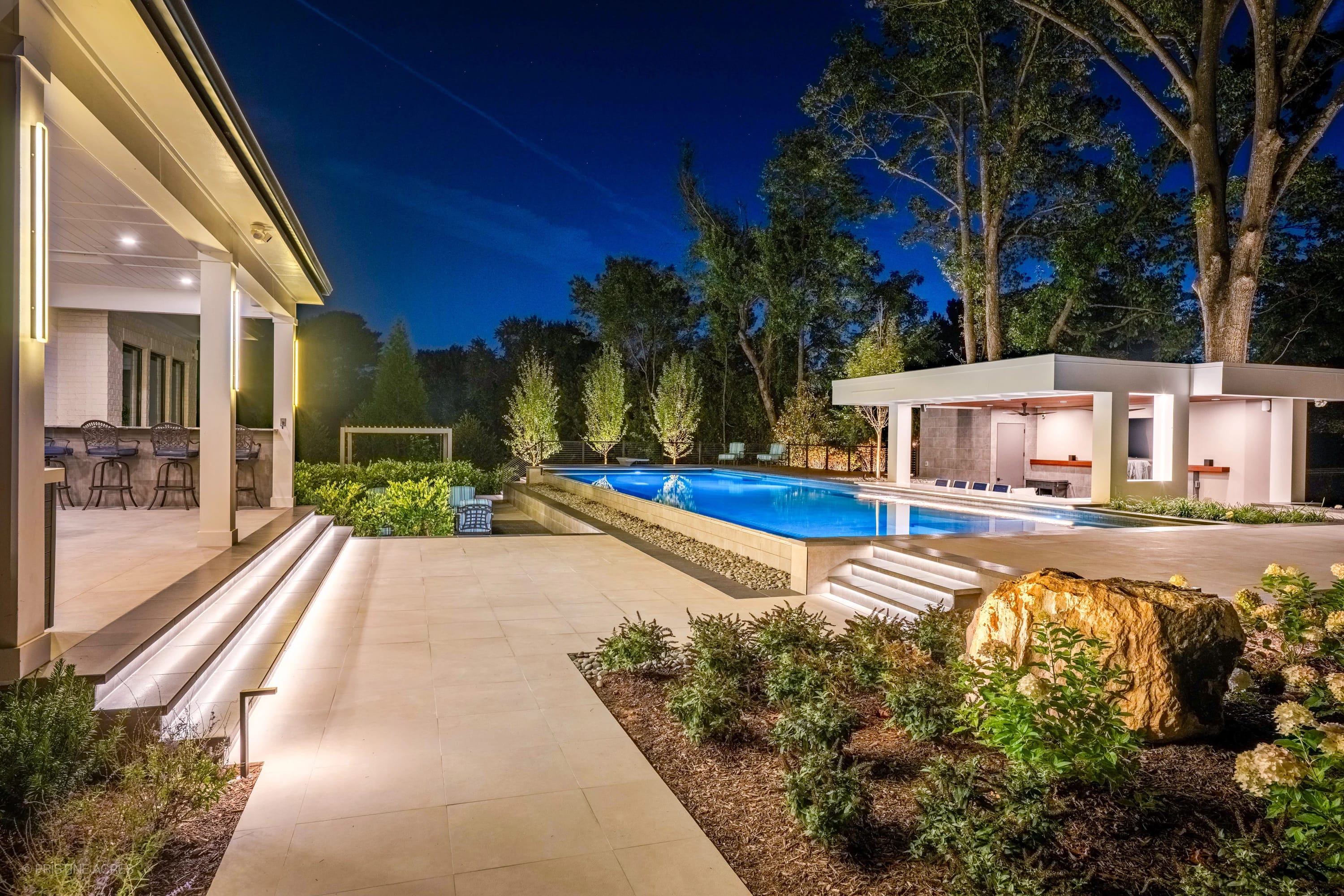 Modern McLean Pool Project in McLean, VA features Black Basalt and Nexus White Porcelain from STONELUX. A sleek pool with blue water, surrounded by lush greenery, modern architecture, and ambient lighting, creates a luxurious, contemporary outdoor space.