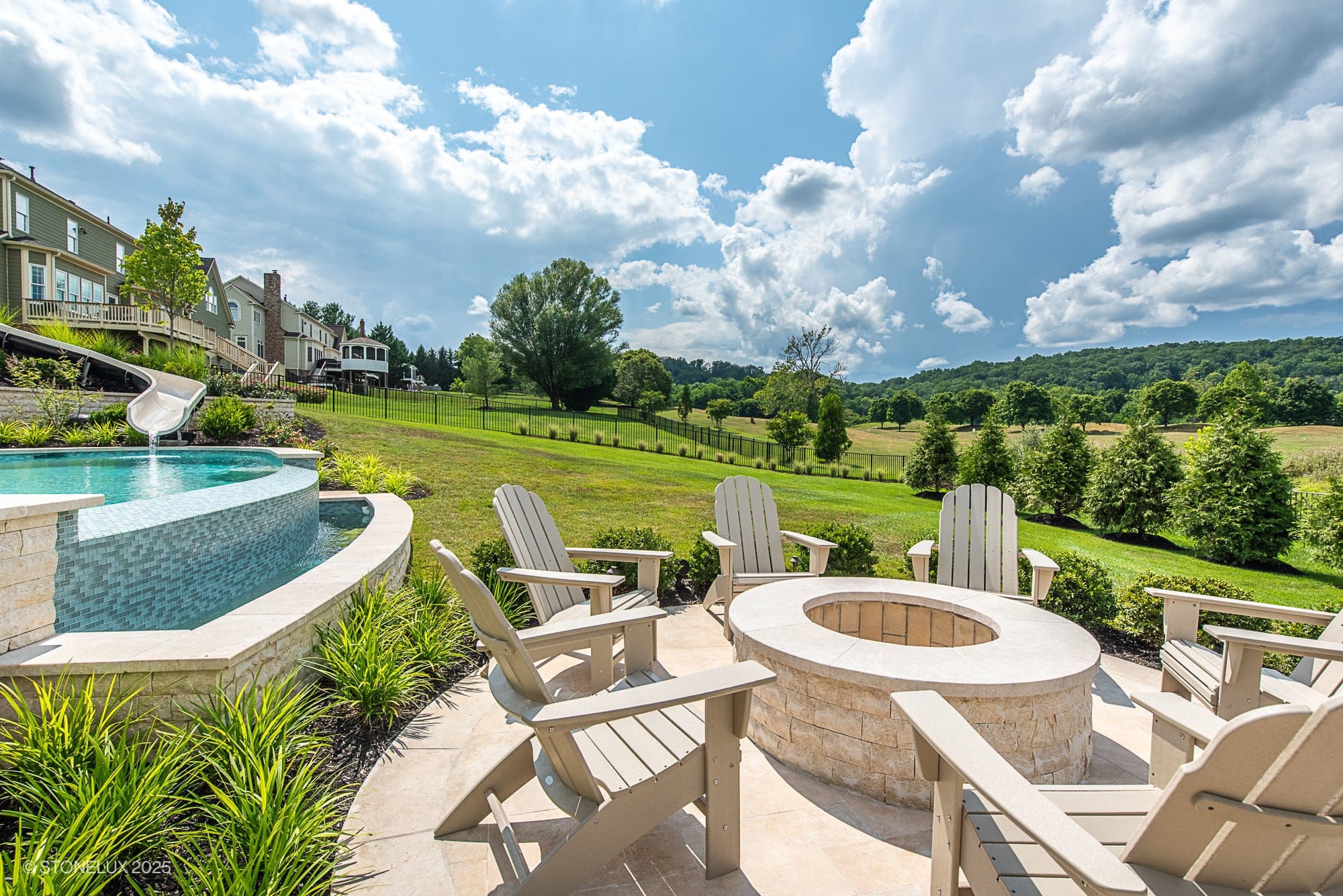 A sunny backyard boasts a Leesburg Grecian Oasis – Ivory Travertine Pool Project, with a circular stone fire pit surrounded by Adirondack chairs, an infinity pool with a slide, lush lawn, trees, and rolling hills under a partly cloudy sky.