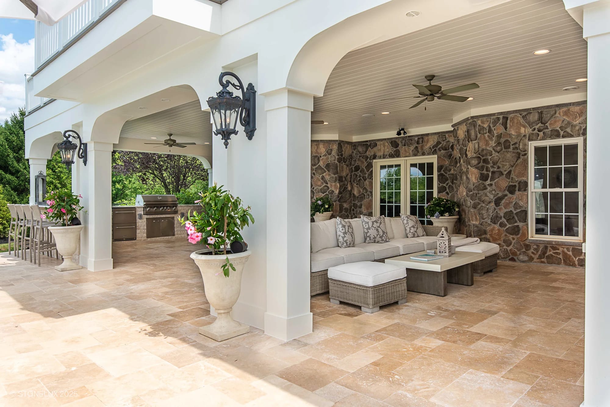 Covered outdoor patio with stone walls, ceiling fans, and a large sectional sofa around a coffee table. Inspired by the Leesburg Grecian Oasis – Ivory Travertine Pool Project, it features a built-in grill, bar area with stools, sconces, and planters.
