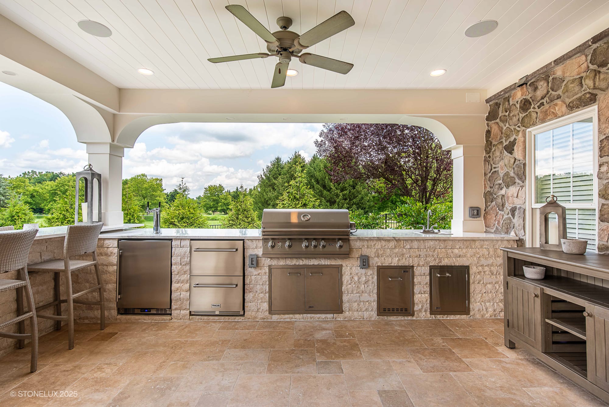 Outdoor kitchen with a built-in stainless steel grill, appliances, and cabinets under a covered patio with stone accents, ceiling fan, bar seating, and scenic views—part of the Leesburg Grecian Oasis – Ivory Travertine Pool Project.