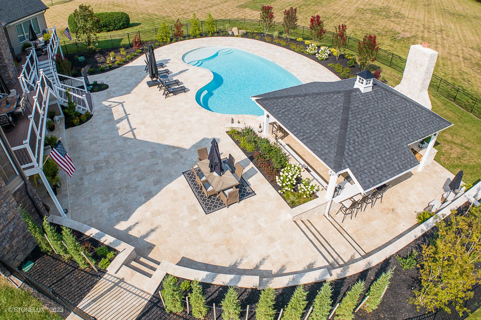 Aerial view of a backyard featuring Beige Bliss: a luxurious travertine pool and pool house with a bar area, surrounded by lounge chairs, landscaped greenery, a patio, fencing, and open grass.