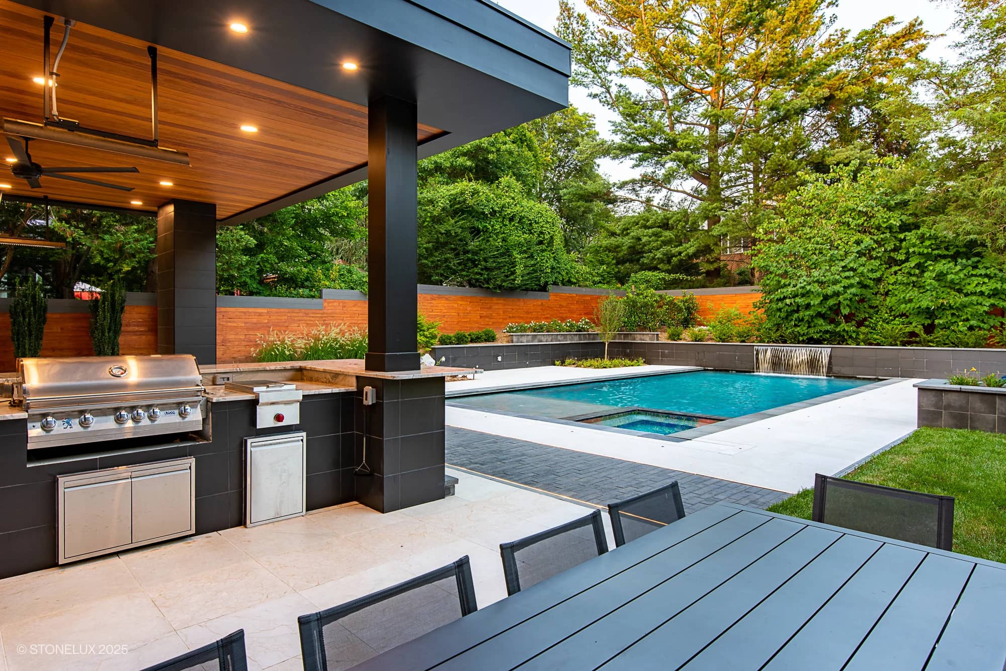 McLean Modern Zen – Outdoor Living in Pera Cream Marble & Black Basalt: Modern backyard with a covered outdoor kitchen and grill area, sleek dining table, and a swimming pool surrounded by greenery and a wooden privacy fence.