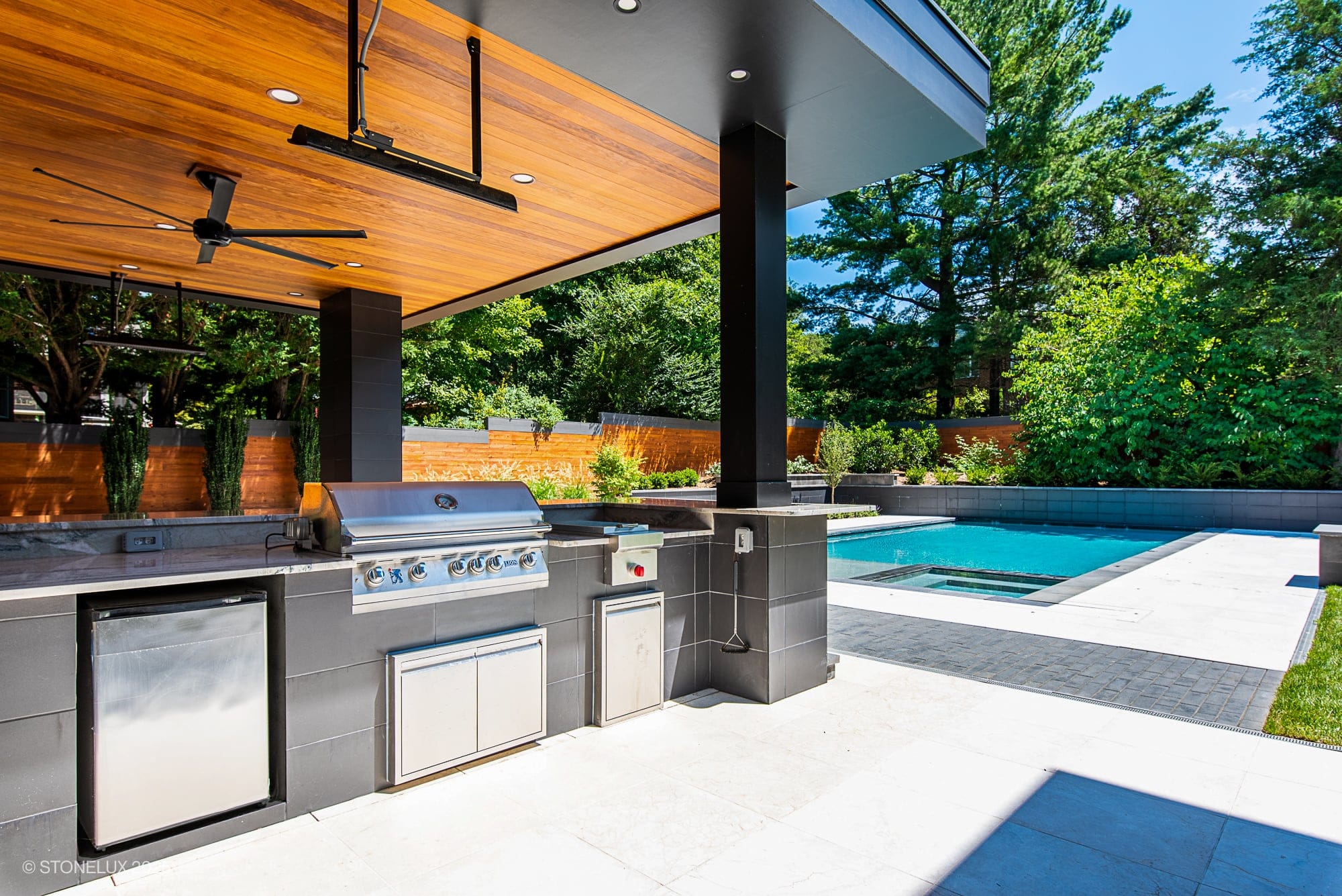 Modern outdoor kitchen with stainless steel grill and sink under a wood-paneled ceiling, set beside a patio featuring McLean Modern Zen – Outdoor Living in Pera Cream Marble & Black Basalt, overlooking a swimming pool surrounded by lush greenery.