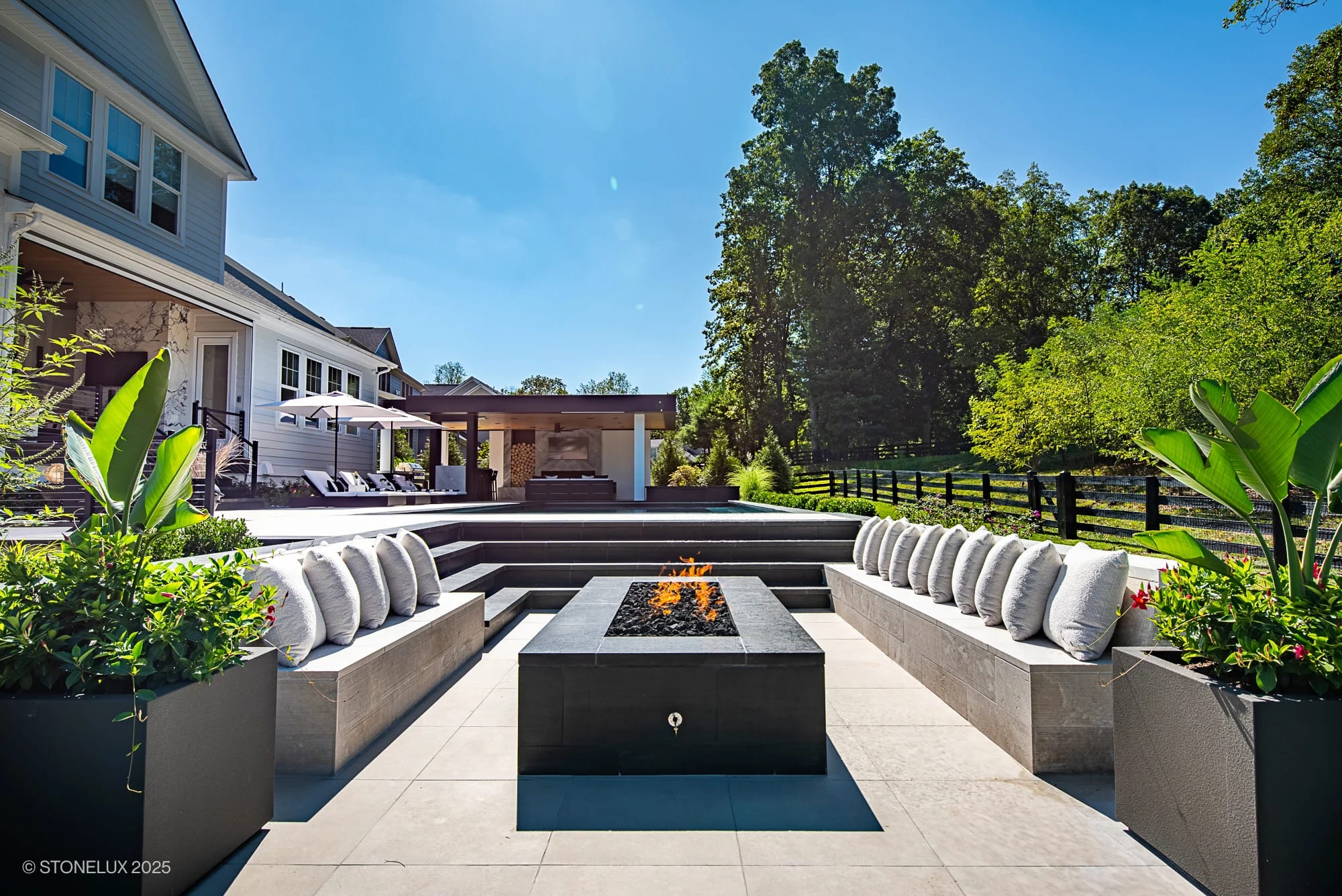 Experience the Aldie Modern Escape – Porcelain & Basalt Luxury Pool Pavilion: a modern outdoor patio with a rectangular fire pit, cushioned seating, stylish planters, sun loungers, and lush greenery set against a stunning contemporary home.