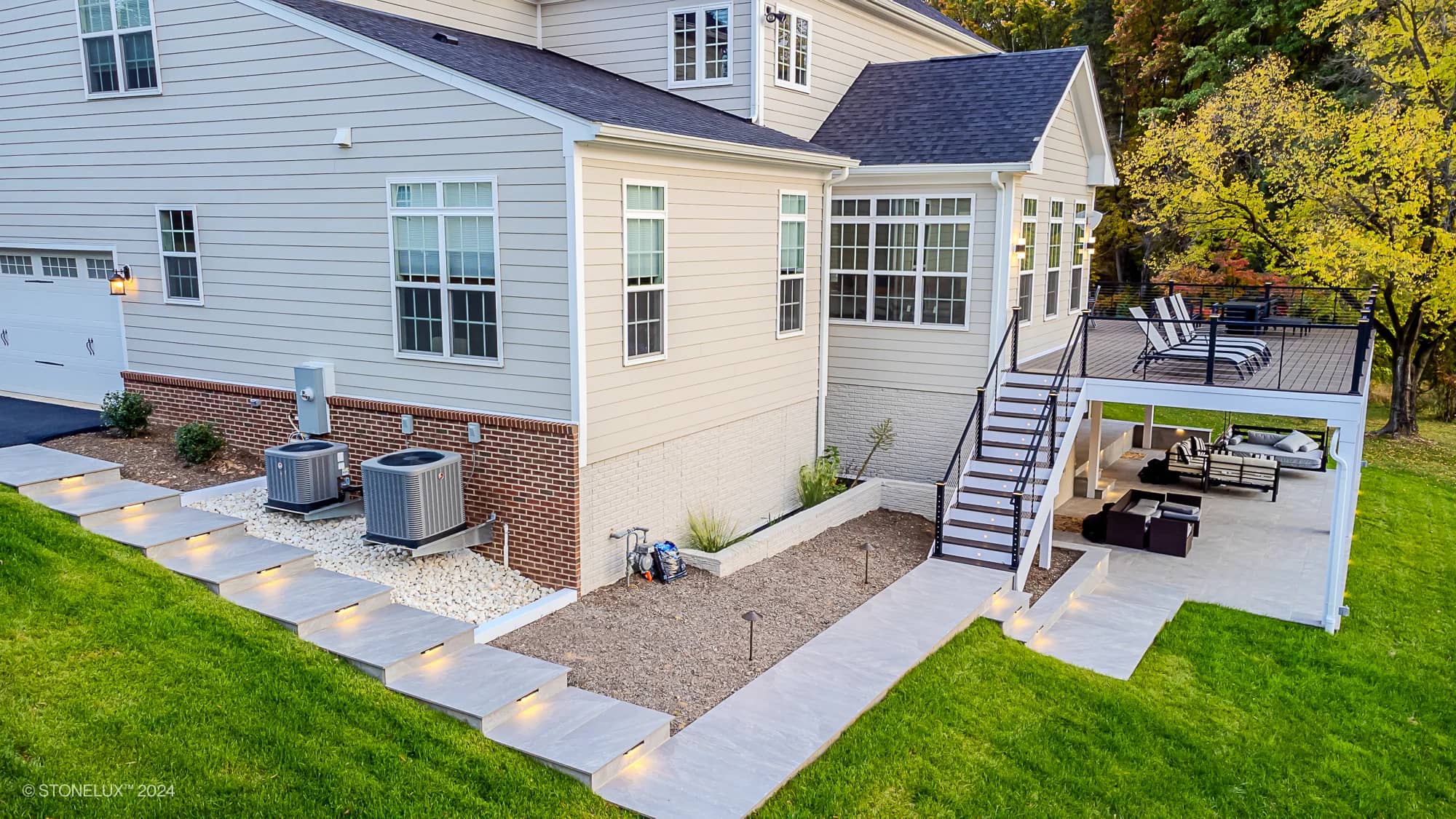 A modern house with a spacious Rasa Grey: Porcelain Paver Patio in Fairfax, upper deck with lounge chairs, and lower seating area. Concrete steps with lights lead to the landscaped yard featuring stones and two HVAC units.