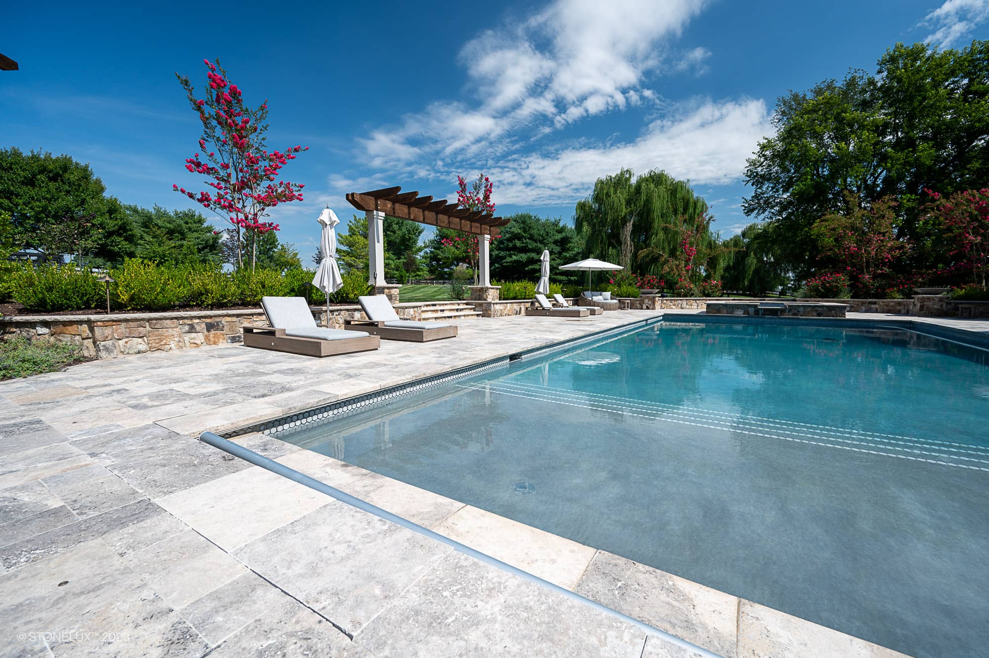A clear outdoor swimming pool with stone decking is the centerpiece of the Silver Expanse: The Silver Travertine Pool Landscape, surrounded by lounge chairs, umbrellas, lush green trees, and flowering plants under a bright blue sky.