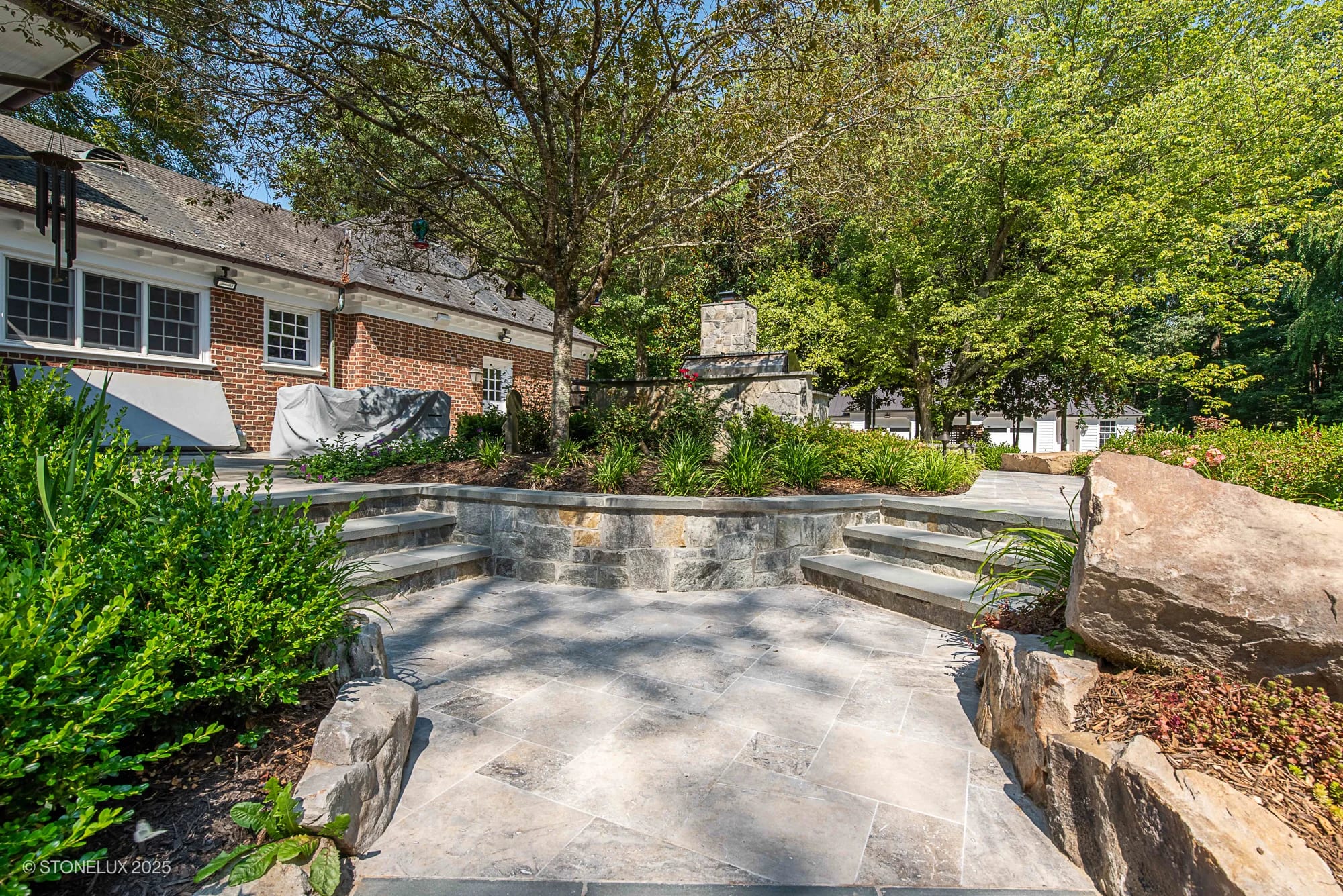 A stone patio, part of the Clifton Cascade Retreat – Silver Travertine Oasis, features curved steps and lush greenery leading to a fire pit area, with a brick house on the left and shade provided by surrounding trees.