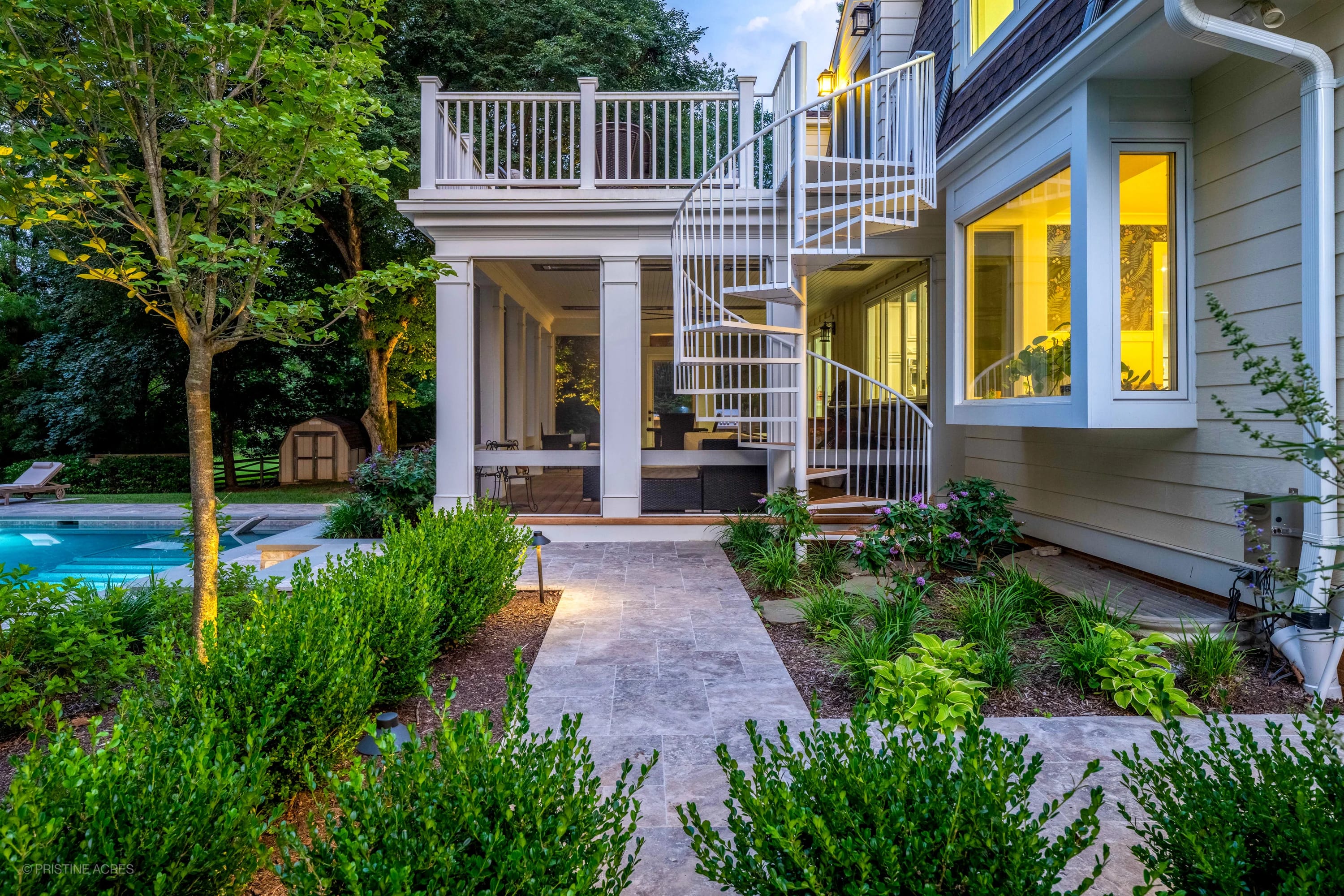 Elegant Vienna Pool Retreat in Vienna, VA showcases Silver Travertine paths leading to a serene pool. The lush greenery and modern spiral staircase create a tranquil atmosphere. Bluestone accents and warm lighting enhance this luxurious outdoor oasis.