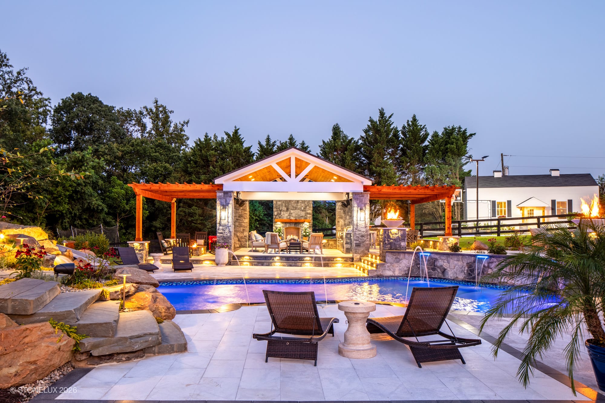 Luxurious backyard oasis in McLean, VA featuring Tundra Grey Marble and Black Basalt. Modern pool with blue water, Afyon White Marble accents, surrounded by lush greenery, elegant pergola, and ambient lighting, creating a serene, upscale atmosphere.