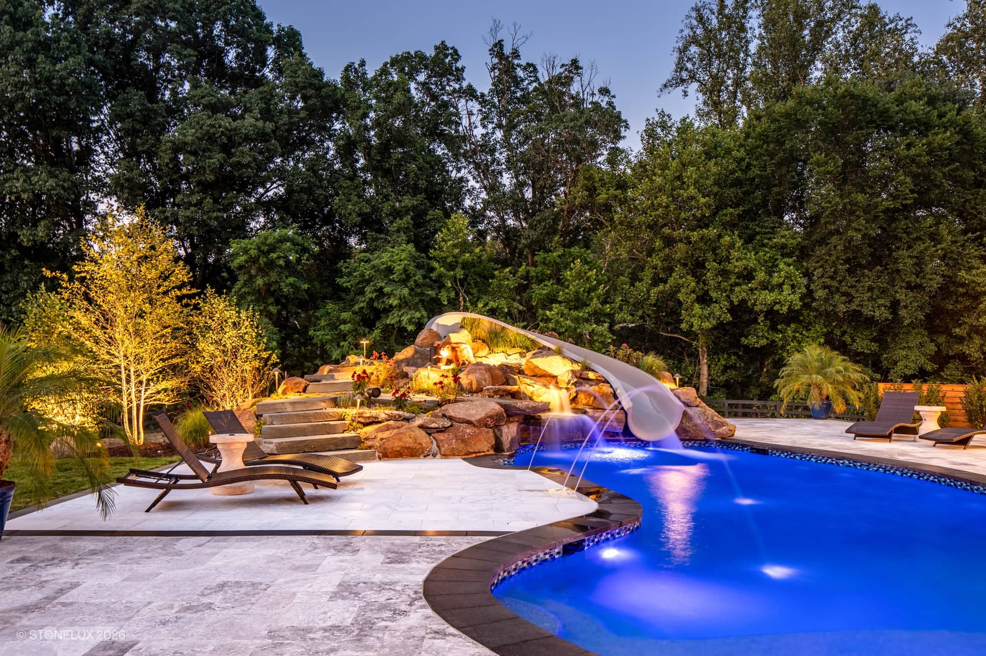 Luxurious backyard oasis in McLean, VA featuring Tundra Grey Marble and Black Basalt pavers. The modern design includes a sparkling blue pool, elegant Afyon White Marble accents, lush landscaping, lounge chairs, and ambient lighting for a serene atmosp...
