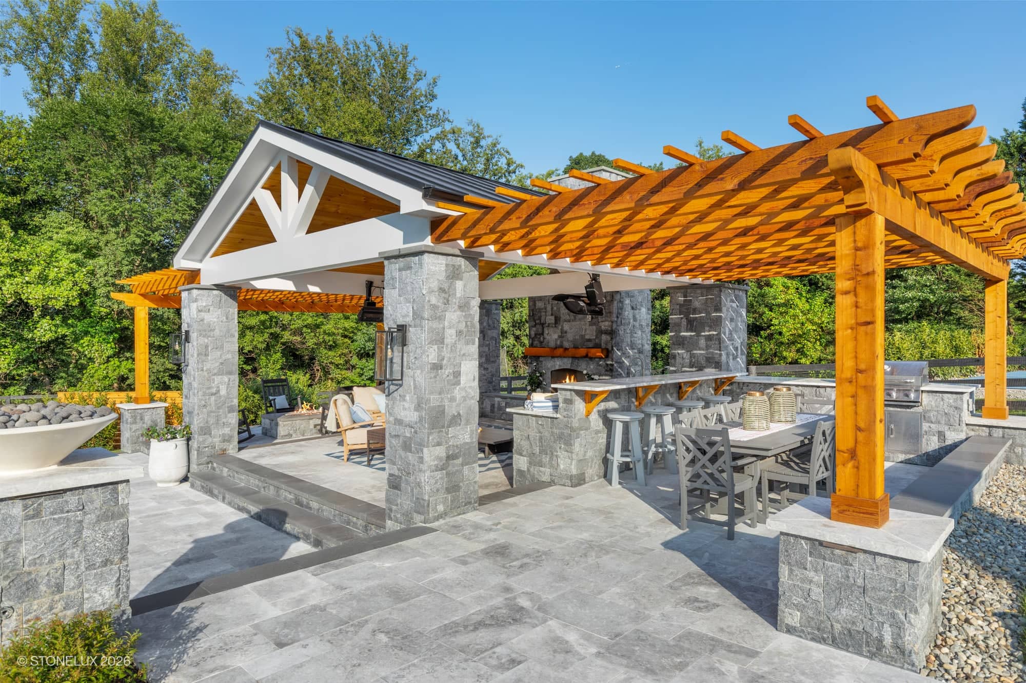 Backyard oasis in McLean, VA featuring Tundra Grey Marble pavers and Black Basalt pillars. Modern design includes a pergola, outdoor kitchen, and seating area. Surrounded by lush greenery, the space offers a serene, luxurious atmosphere for relaxation.