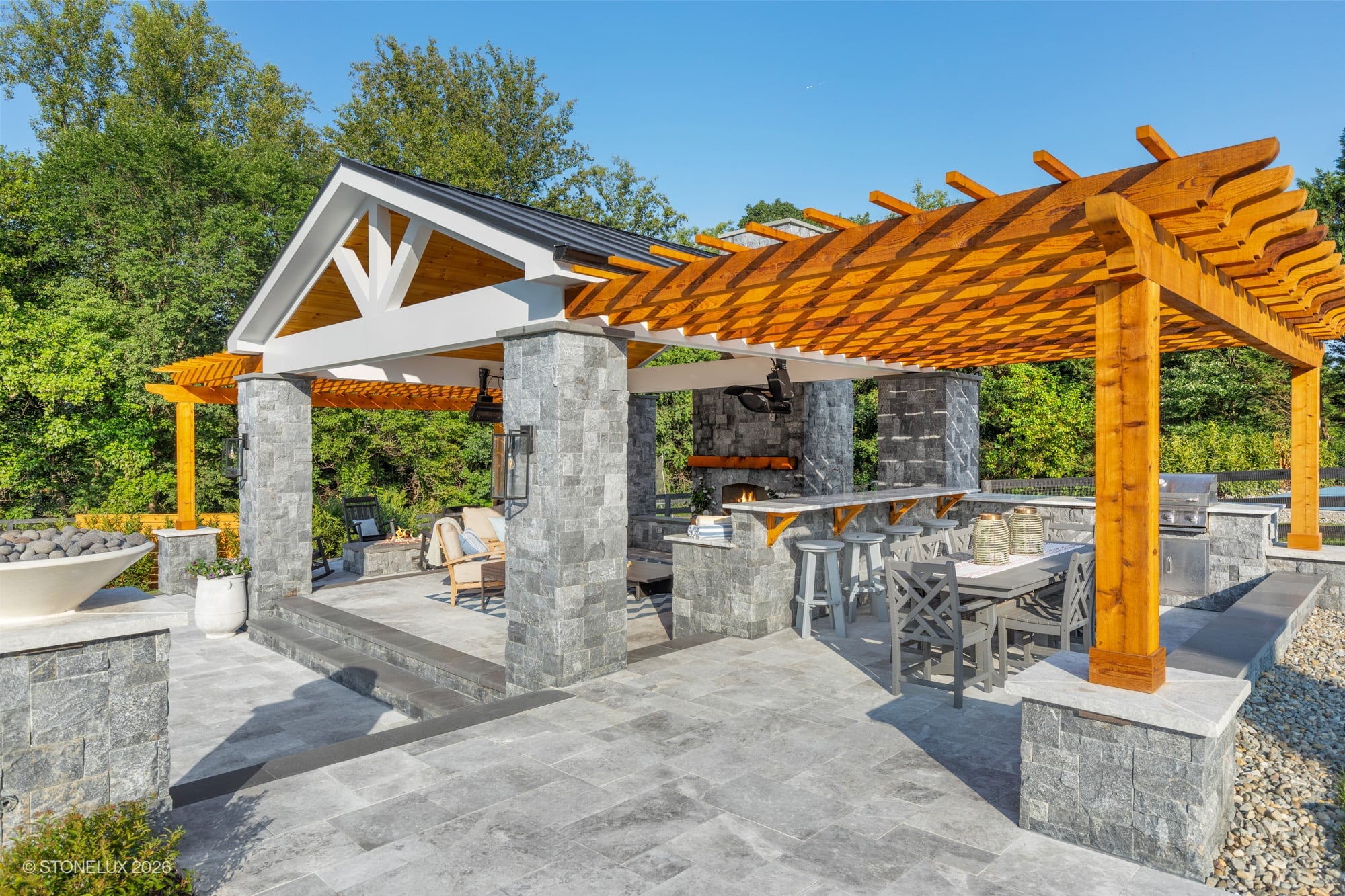 Backyard oasis in McLean, VA featuring Tundra Grey Marble pavers and Black Basalt pillars. Modern design includes a pergola, outdoor kitchen, and seating area. Surrounded by lush greenery, the space offers a serene, luxurious atmosphere for relaxation.