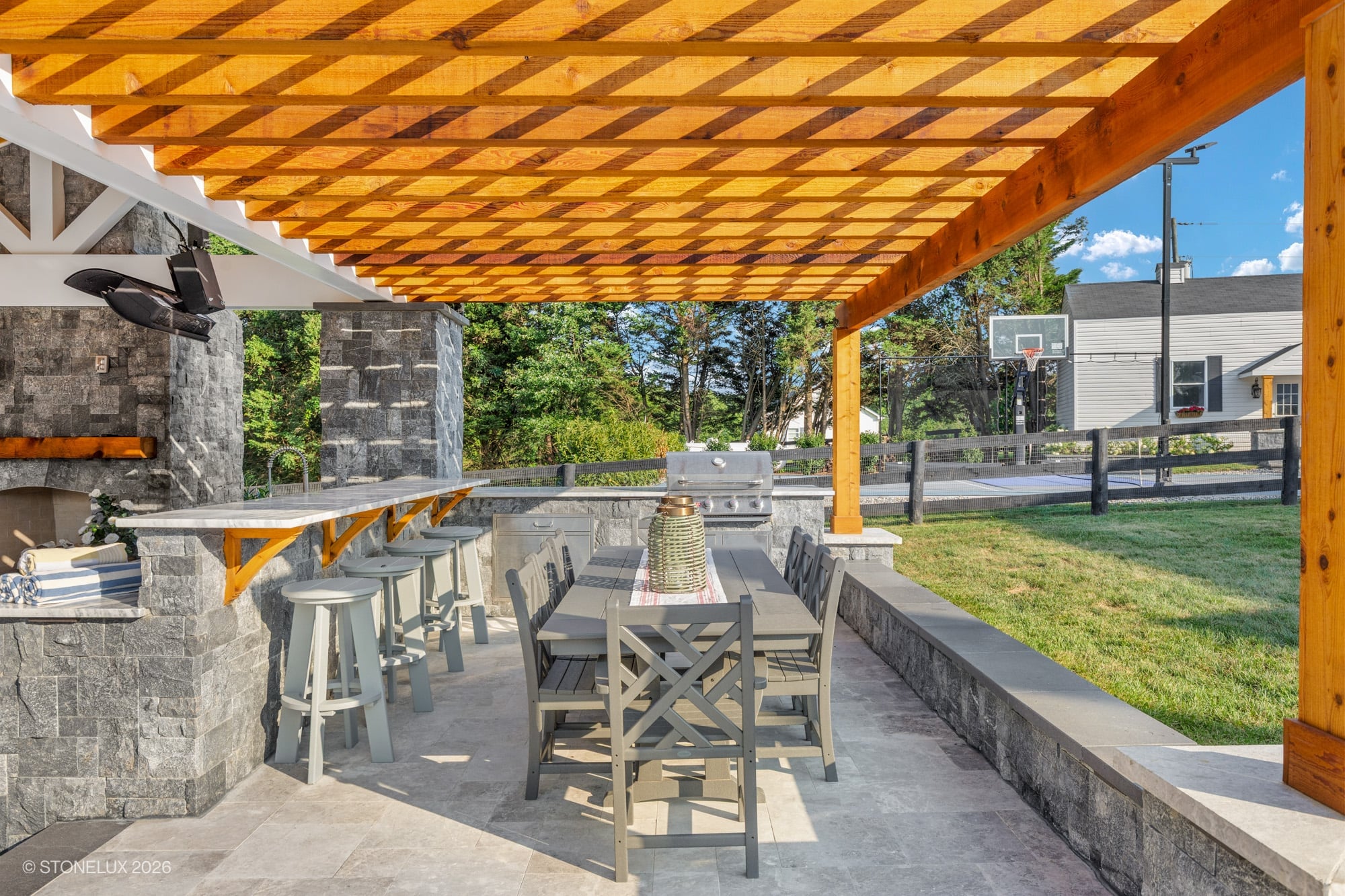 Backyard oasis in McLean, VA featuring Tundra Grey Marble pavers and Black Basalt elements. Modern design with a pergola, outdoor kitchen, and dining area. Lush greenery surrounds the space, creating a serene setting under natural light. Perfect for en...