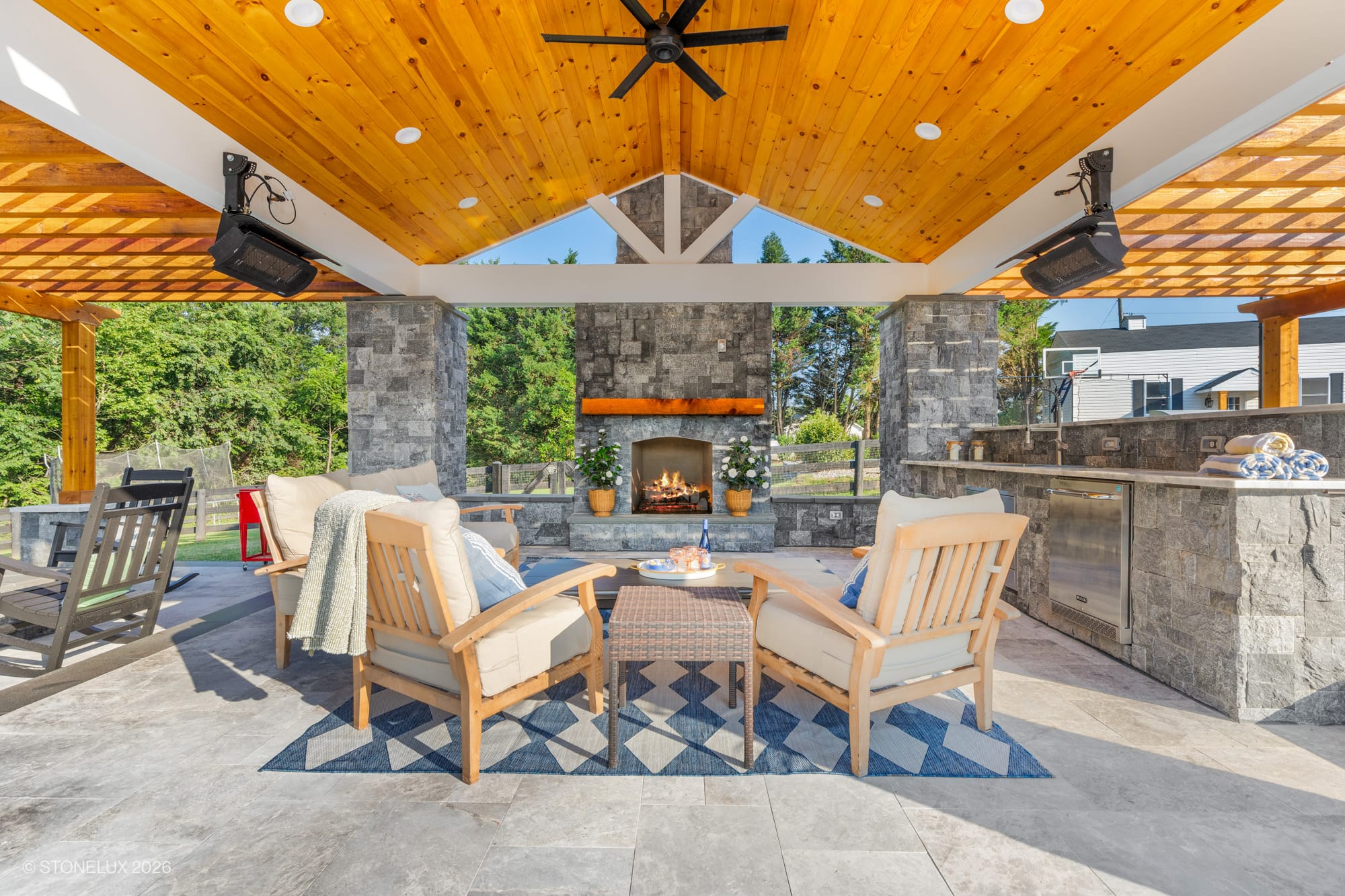 Luxurious backyard oasis in McLean, VA featuring Tundra Grey Marble flooring and Black Basalt accents. Modern patio with wooden ceiling, cozy seating around a stone fireplace, and lush landscaping. Perfect blend of natural stone and contemporary design.