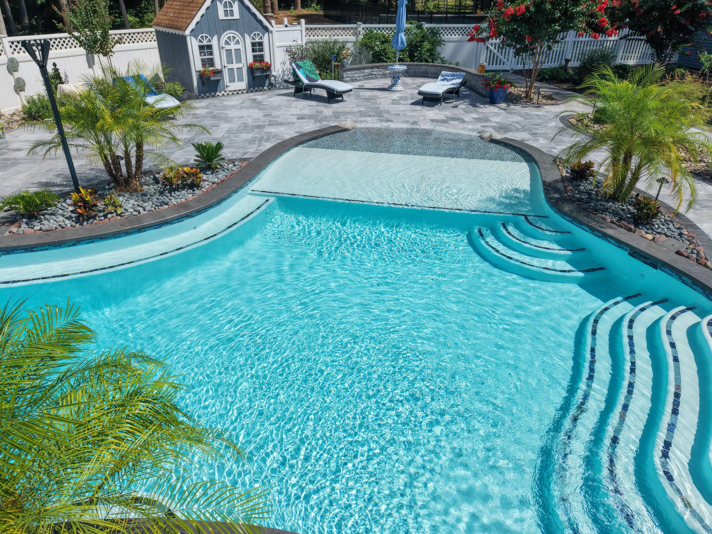 Aegean Grey Marble Pool Retreat in Salisbury, MD, features sleek black basalt coping. The modern design includes clear blue water, curved steps, and lush tropical landscaping. The grey patio harmonizes with white loungers and vibrant plants, creating a...
