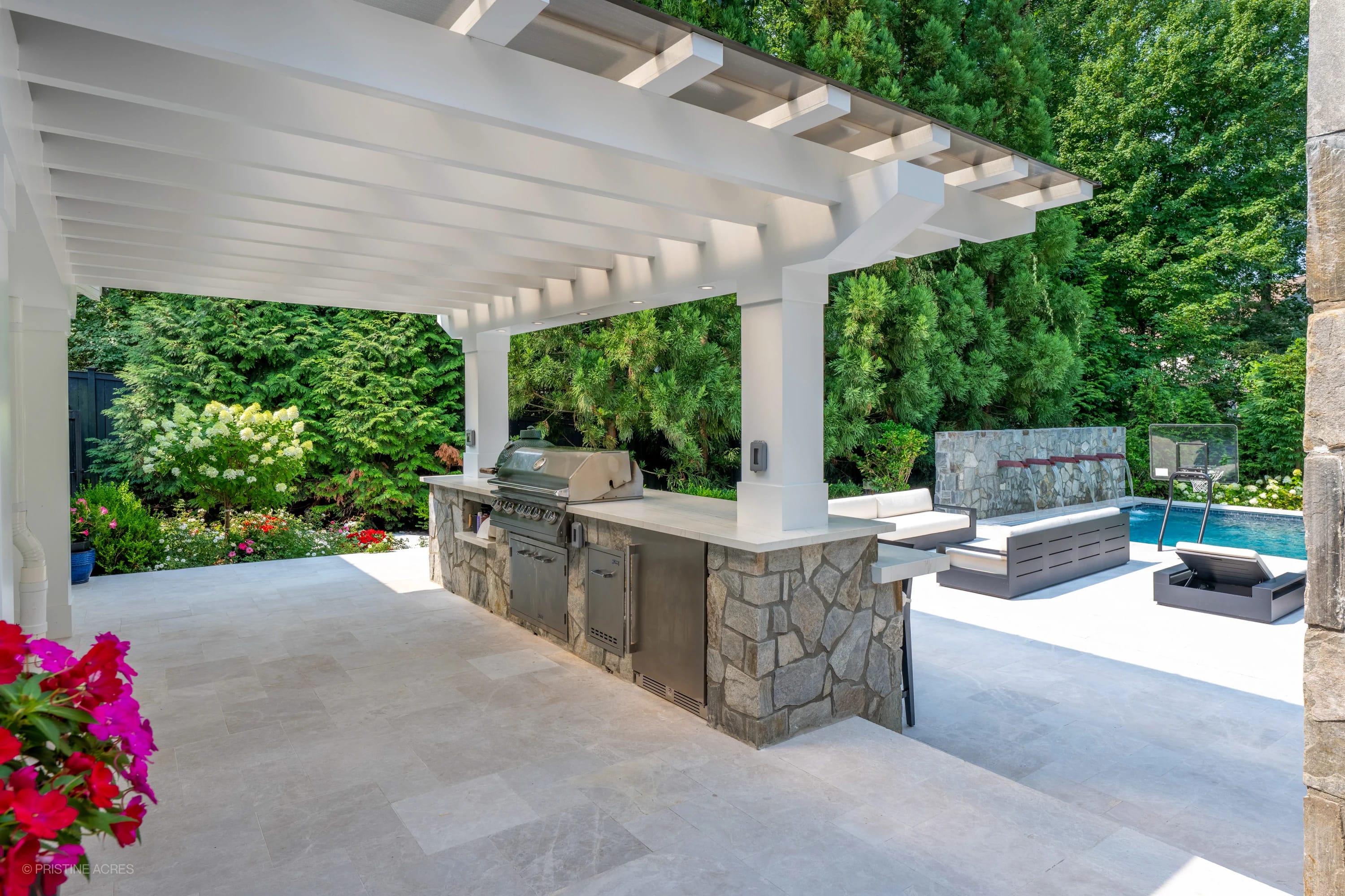 Outdoor living space in McLean, VA, featuring Pera Cream Marble flooring. The modern design includes a stainless steel grill under a pergola, surrounded by lush greenery. Adjacent is a pool with blue water, stone accents, and contemporary seating, crea...