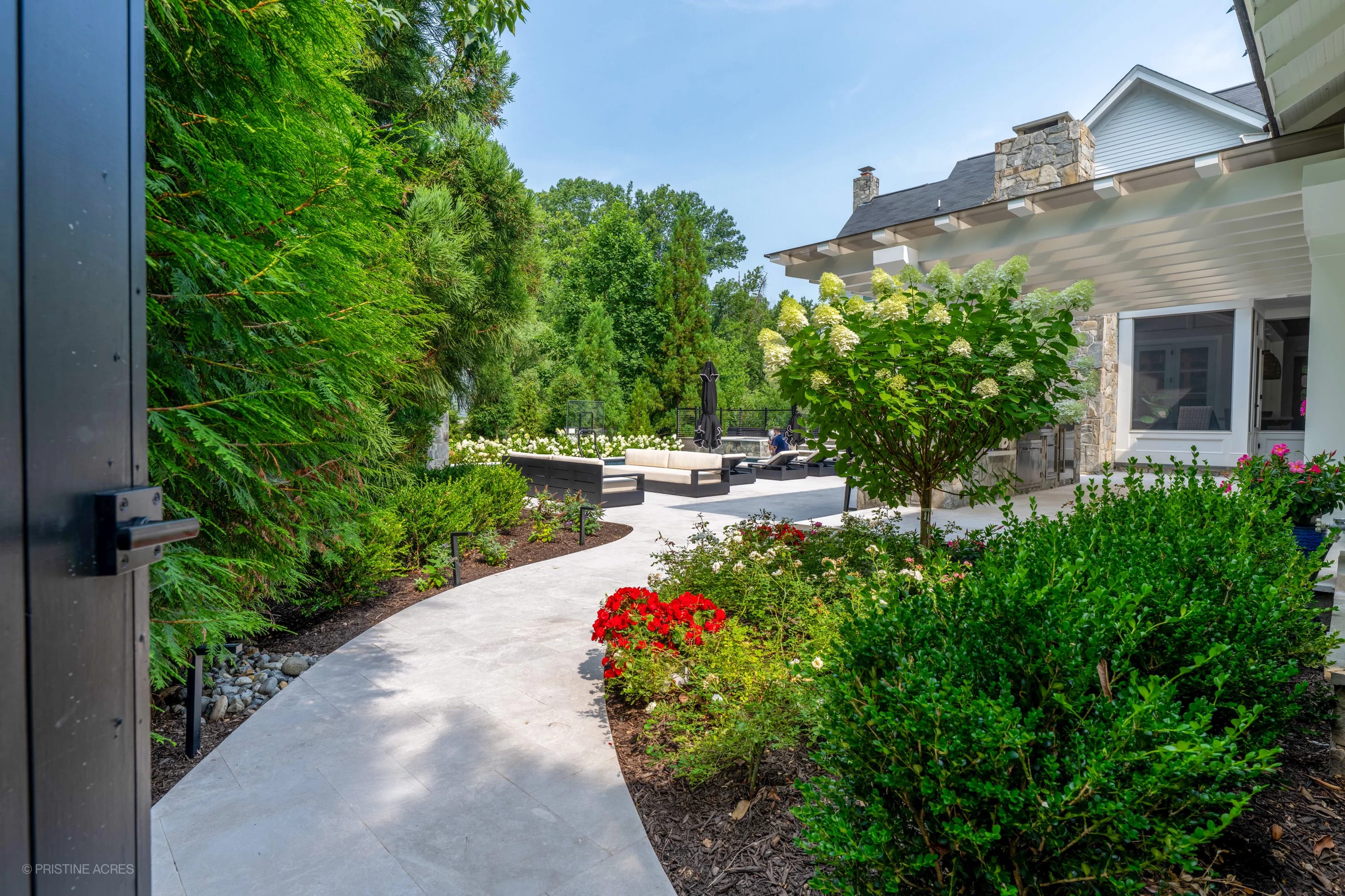 Luxurious outdoor setting in McLean, VA featuring Pera Cream Marble pavers. The elegant patio is surrounded by vibrant greenery, colorful flowers, and modern outdoor furniture. A stone facade house complements the Mediterranean-inspired design.