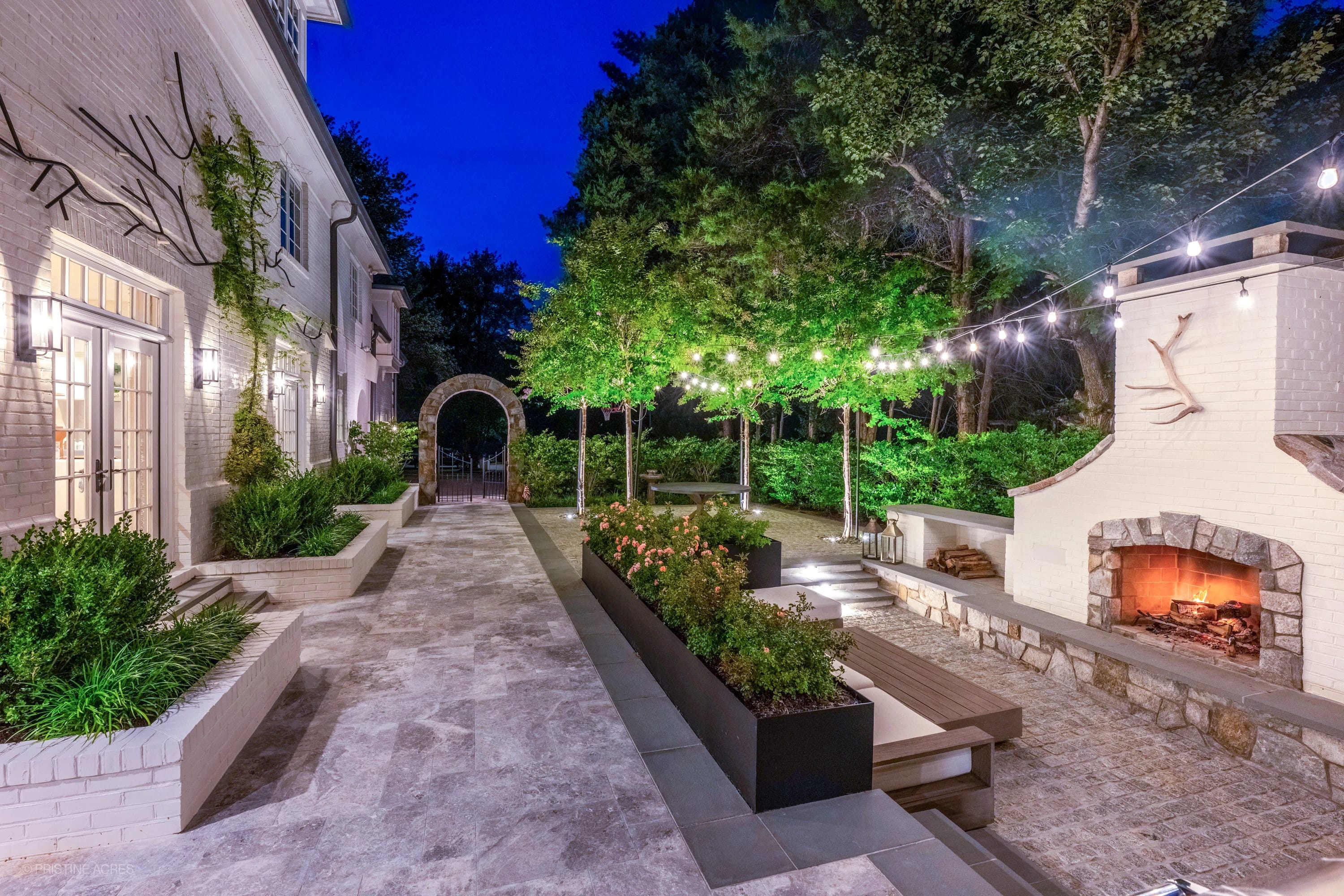 Tundra Grey Marble pool deck in McLean, VA features elegant bluestone coping. Surrounded by lush greenery, string lights, and a cozy outdoor fireplace, this modern patio exudes warmth and sophistication, perfect for luxurious evening gatherings.