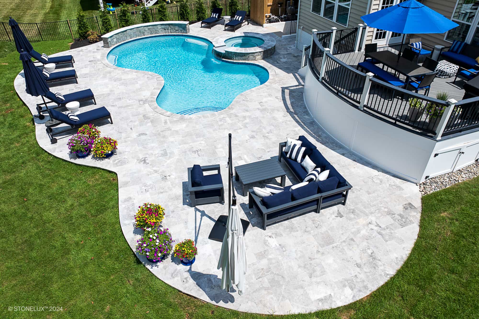 A backyard in Ashburn, VA features a curvy swimming pool with Tundra Grey Marble accents, an attached hot tub, lounge chairs, navy-cushioned seating areas, potted flowers, and a patio with a blue umbrella near the house.