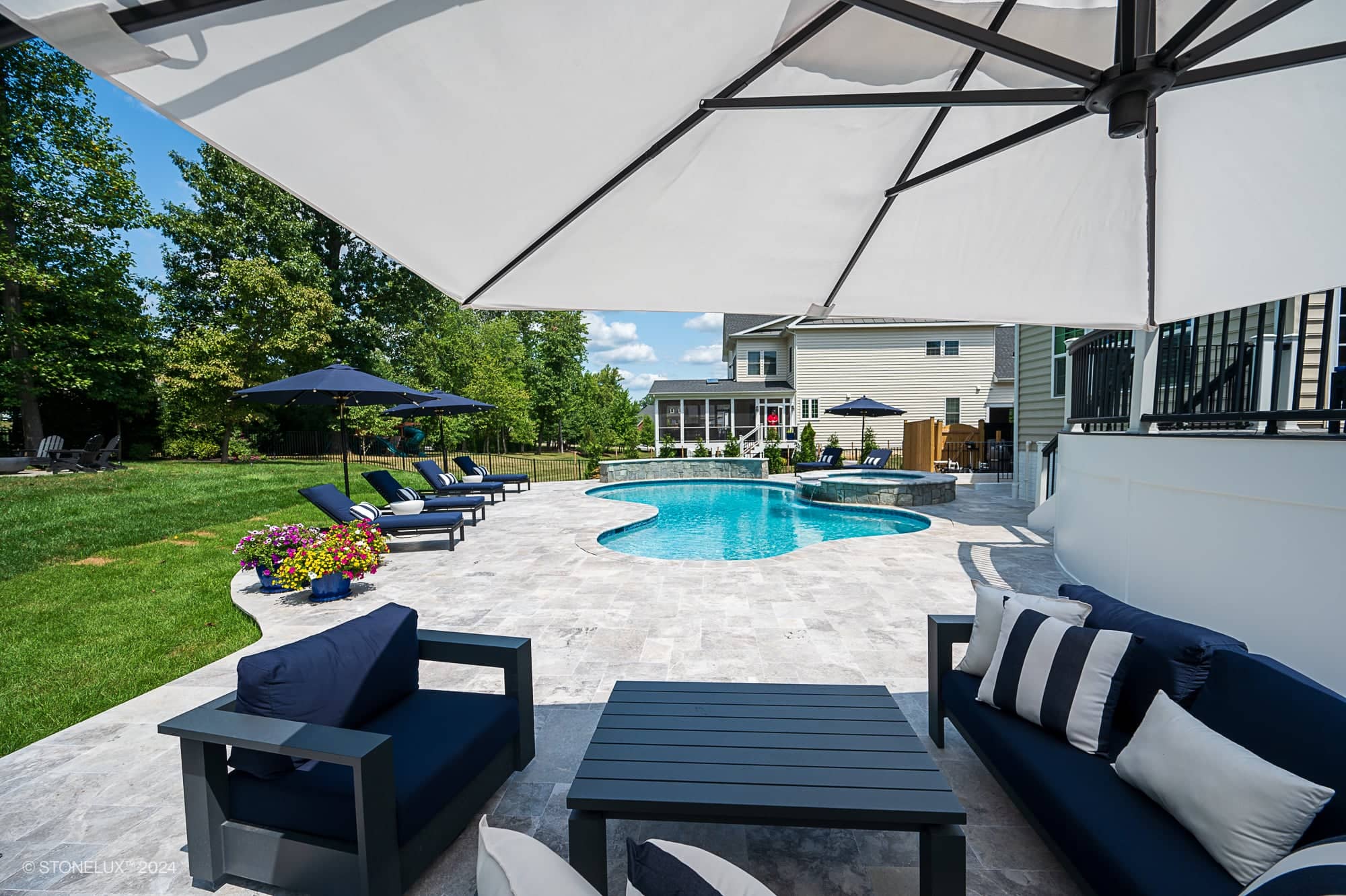 A backyard patio with modern outdoor furniture under a large umbrella overlooks a kidney-shaped swimming pool featuring Tundra Grey Marble. Lush green lawn, trees, and a light-colored house complete this pool and patio project in Ashburn VA.