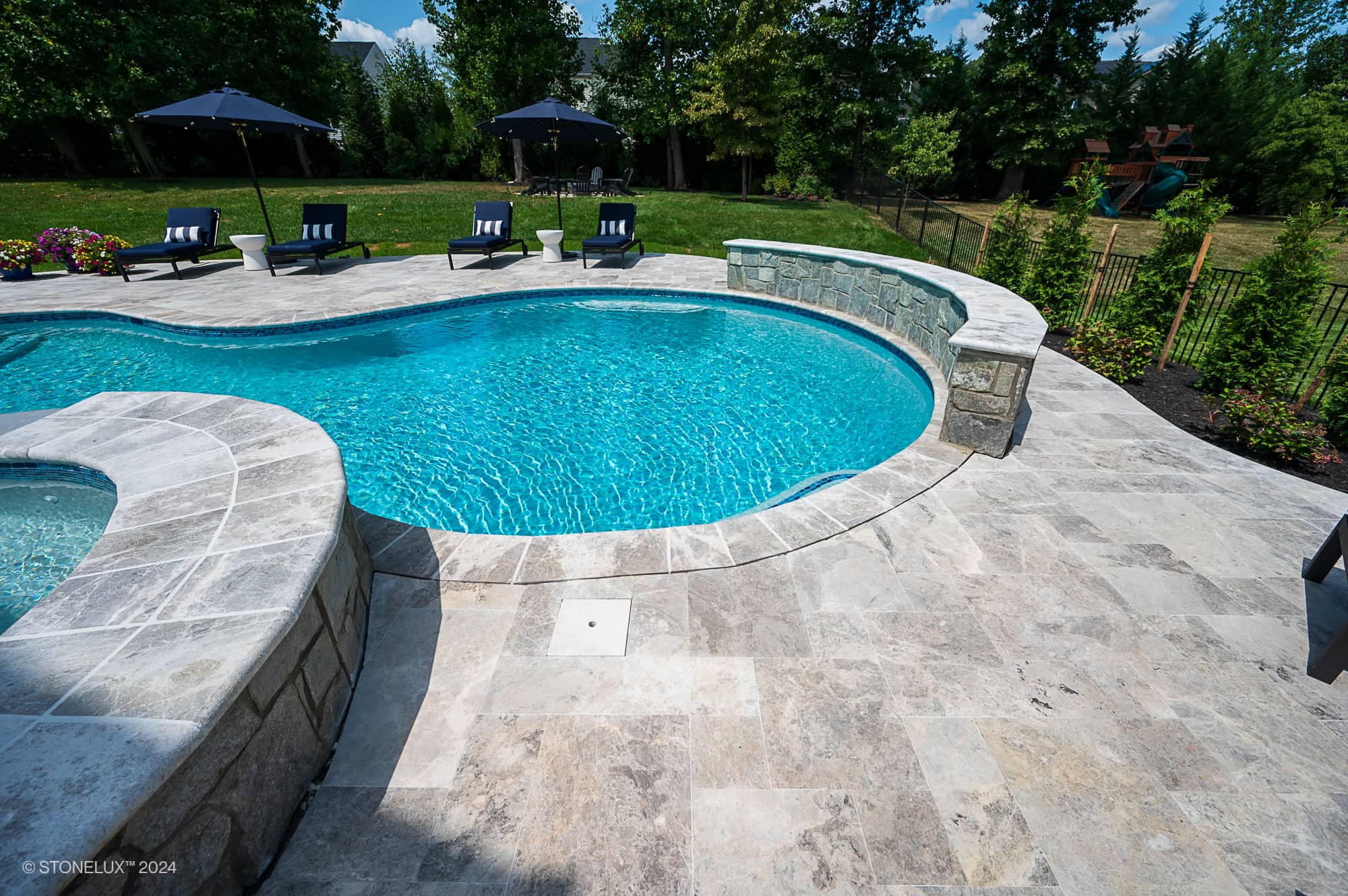 A backyard swimming pool with clear blue water is surrounded by Tundra Grey Marble stone tile decking. Lounge chairs and umbrellas sit on the grass nearby, with trees and a playset visible in this Pool and Patio Project in Ashburn VA.