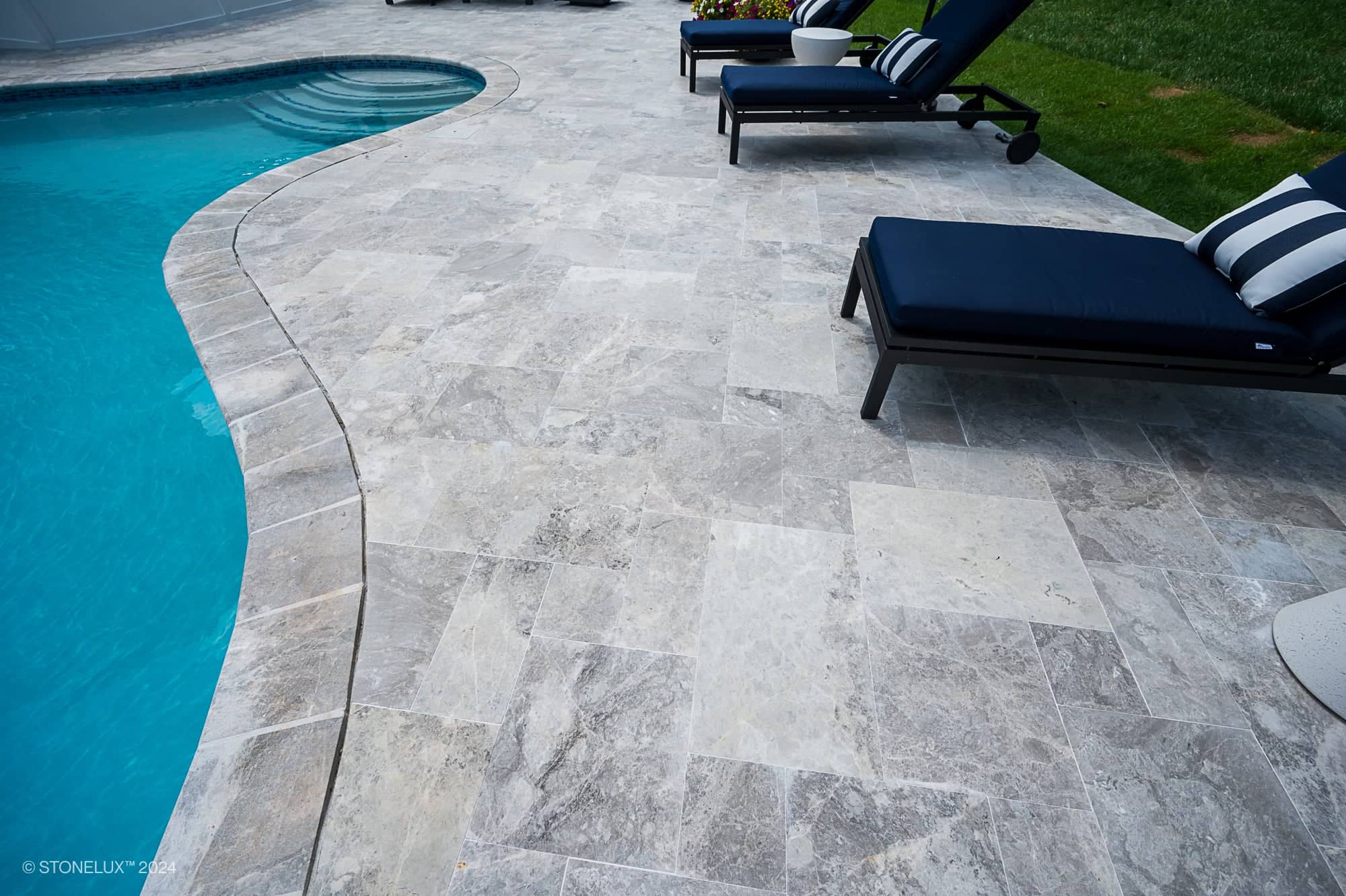 Curved-edge swimming pool with Tundra Grey Marble tile deck, two navy blue lounge chairs with striped pillows, and a patch of green grass in the background. Pool and Patio Project in Ashburn VA.