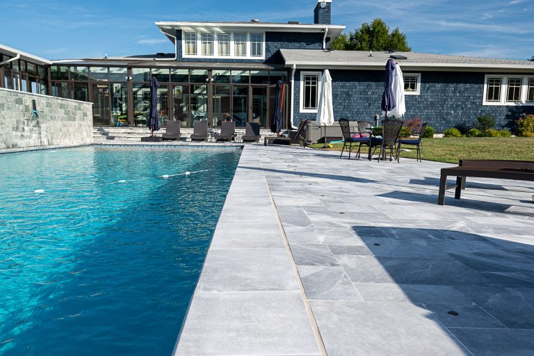 A luxurious backyard featuring a large swimming pool with Arctic Grey Marble Pavers, flanked by a sunken patio area and a modern house with extensive glass walls and stone exteriors under a clear blue sky.