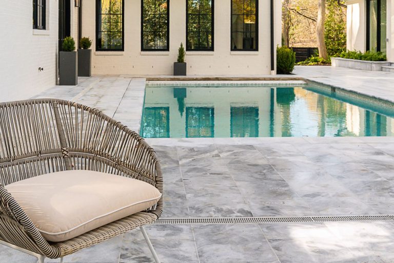 A luxurious backyard featuring a large swimming pool with Arctic Grey Marble Pavers, flanked by a sunken patio area and a modern house with extensive glass walls and stone exteriors under a clear blue sky.