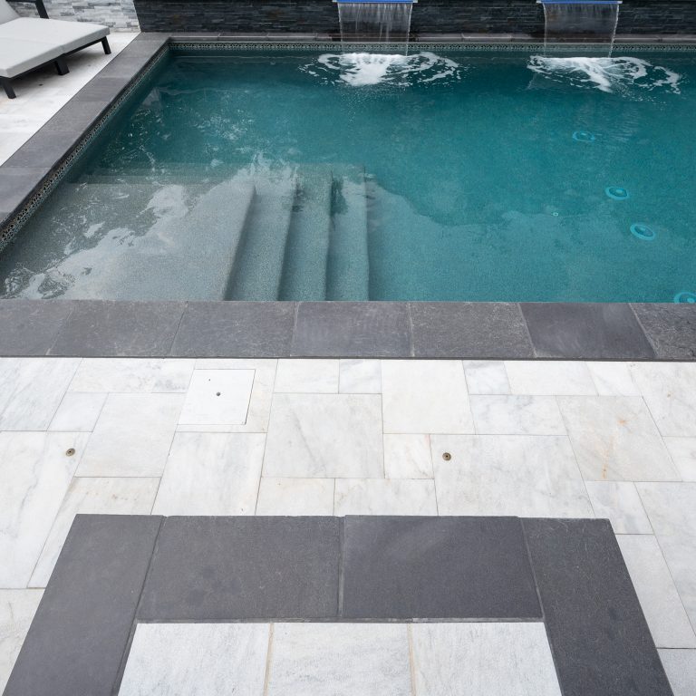 An outdoor swimming pool with stairs visible under the water, surrounded by a tiled deck featuring the Black Basalt Collection design, under a cloudy sky.