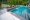 An elegant outdoor swimming pool with clear blue water, surrounded by black basalt pool coping and green foliage, featuring a small waterfall and white picket fence in the background.