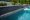 A serene pool with clear blue water featuring three cascading waterfalls along a dark stone wall with black basalt wall caps, bordered by lush greenery and a traditional white picket fence.