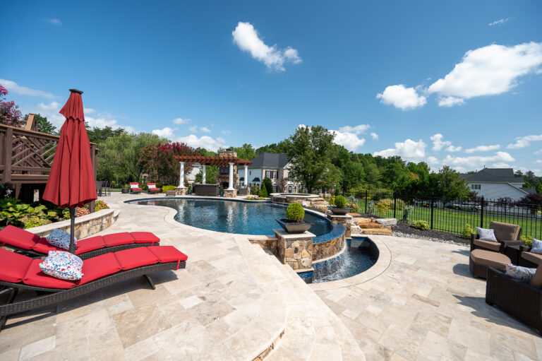 Luxurious backyard with a curvy swimming pool featuring walnut travertine pool copings, stone patio, red lounge chairs, and an outdoor kitchen pavilion under a clear blue sky.
