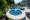 A luxurious backyard featuring a curved swimming pool with an attached circular jacuzzi, surrounded by lush landscaping and Philly Travertine Pavers and Copings, adjacent to a large house.