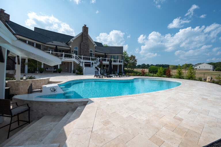A luxurious swimming pool with clear blue water, surrounded by an Ivory Travertine Tumbled French Pattern 3cm Paver - Premium stone patio and two lounge chairs, overlooking a serene pond and landscaped garden under a bright blue sky.