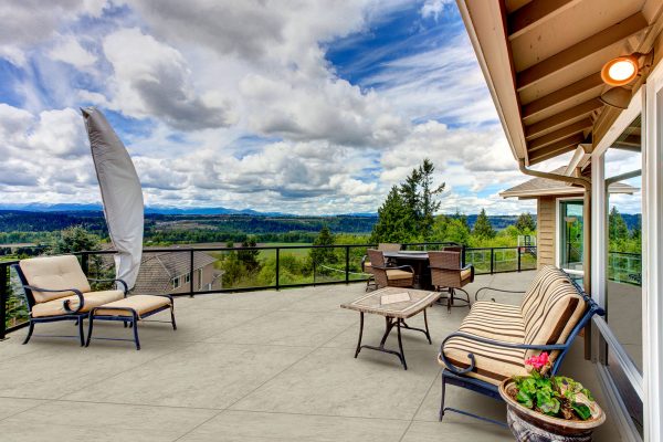 Spacious outdoor patio of a home with modern furniture and Egypt Stone 24"x24" 2CM Matte Rectified Porcelain Paver flooring overlooking a vast, scenic landscape with lush greenery and distant hills under a dramatic cloudy sky.