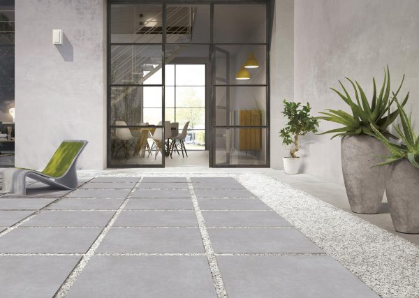 A modern office lobby with large glass doors, Ark Silver 24"x48" 2CM Matte Rectified Porcelain Paver floors, concrete walls, and potted plants. A green chair is on the left, leading into a brightly lit interior workspace.
