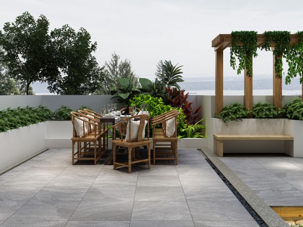 An outdoor dining area on a Rasa Gray 24"x24" 2CM Matte Rectified Porcelain Paver patio overlooking the sea. It features a wooden table set with dinnerware, surrounded by chairs, and flanked by lush greenery and plants.