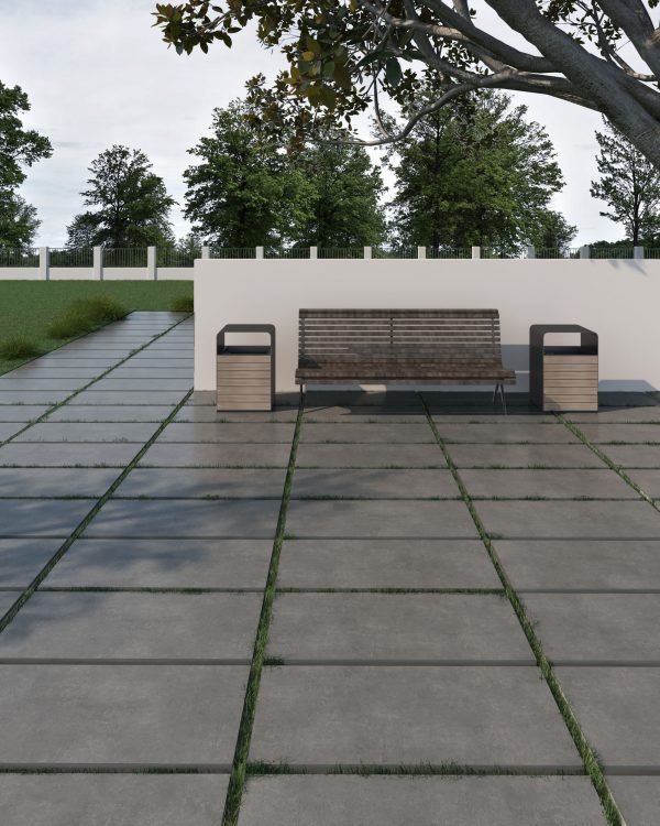 Ark Antracite 24"x24" 2CM Matte Rectified Porcelain Paver park bench with black supports positioned on a rectified porcelain pathway with grass strips, surrounded by trees and a low white wall, under a clear sky.