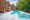 Luxurious backyard with a large swimming pool and a smaller attached hot tub, surrounded by an Afyon White Marble deck. Red umbrellas, lounging chairs, and flowering plants decorate the area next to