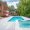 Luxurious backyard with a large swimming pool and a smaller attached hot tub, surrounded by an Afyon White Marble deck. Red umbrellas, lounging chairs, and flowering plants decorate the area next to