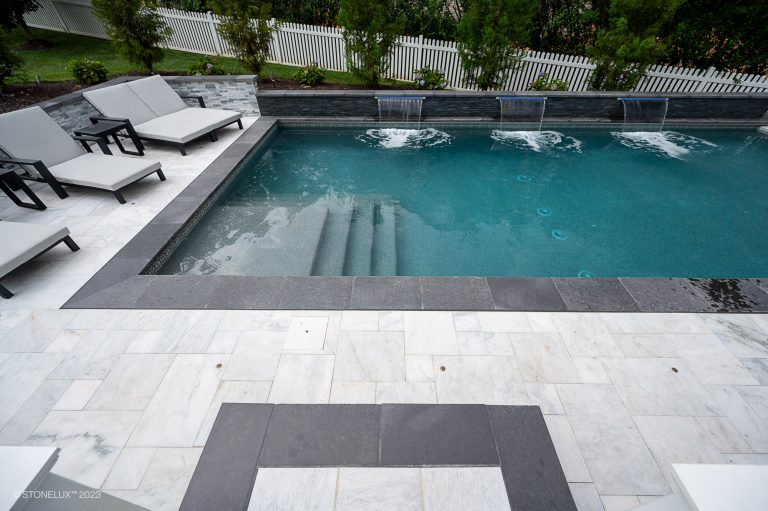 A serene backyard pool with Black Basalt Brushed Eased Edge Pool copings, surrounded by white fencing, lush greenery, and a dark stone waterfall feature. Black basalt veneers and stone tiles complete the tranquil setting.
