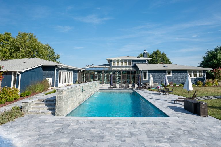 A luxurious backyard with a large swimming pool, surrounded by Arctic Grey Marble Pavers. Adjacent are modern, gray stone buildings and a lush garden under a clear blue sky.