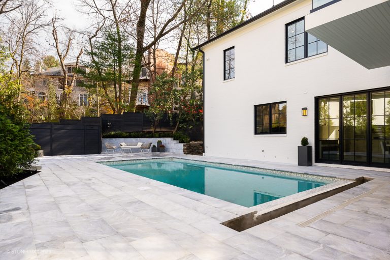 A modern backyard with a rectangular swimming pool, surrounded by Arctic Grey Marble pavers and copings. A white two-story house with black windows is to the left, and outdoor seating is visible.