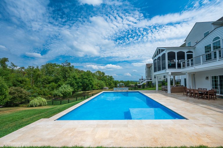 A luxurious backyard featuring a rectangular swimming pool, surrounded by ivory travertine pavers and copings, and a large white house with multiple balconies set against a backdrop of a lush green landscape.