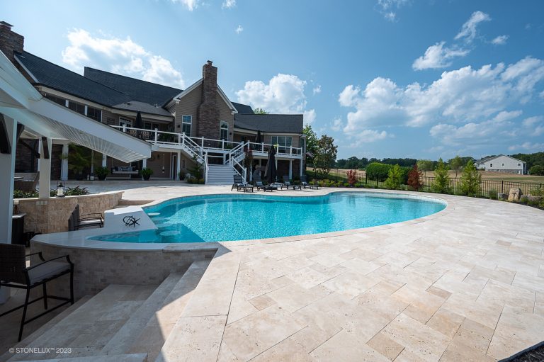A luxurious backyard with a large, curving swimming pool next to a spacious house, featuring an ivory travertine French pattern stone patio and a covered outdoor dining area under clear skies.