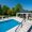 An elegant backyard featuring a rectangular swimming pool, surrounded by spacious Pera Cream Marble Steps with loungers and a gazebo that houses an outdoor seating area. Lush trees border the area under a clear