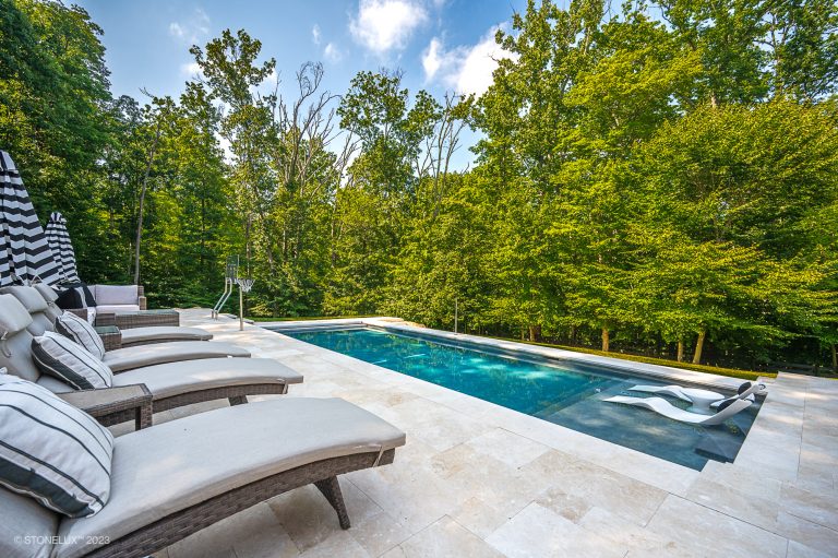 An outdoor swimming pool surrounded by dense green trees, with white lounge chairs on Pera Cream Marble Pavers under a clear blue sky.